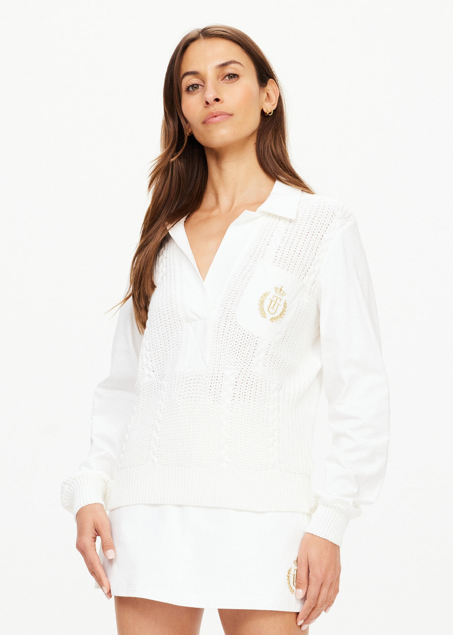 A woman stands confidently against a white background, dressed in an all-white ensemble that includes the Baccarat Violette Sweater from The Upside and a short skirt. The vintage organic cotton sweater is adorned with an embroidered emblem on the chest. Her long brown hair cascades down as she poses with one hand by her side.