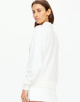 A woman with long brown hair is standing sideways, looking at the camera. She is wearing a Baccarat Violette Sweater by The Upside, and shorts crafted from organic cotton, set against a plain white background.