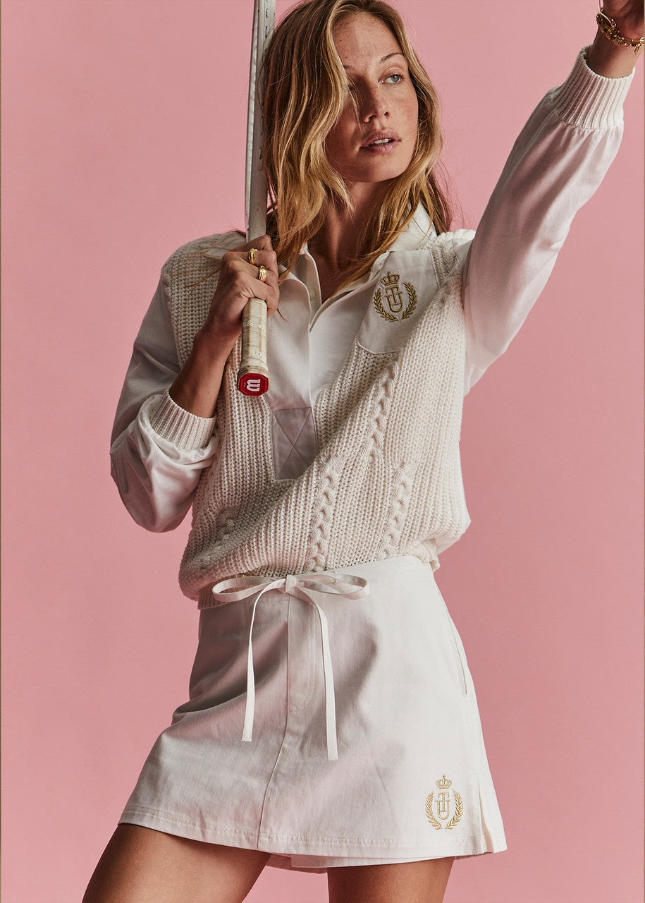 A woman holding a tennis racket poses against a pink background. She wears a relaxed silhouette featuring The Upside's Baccarat Violette Sweater, made from organic cotton, over a white shirt and an embroidered skirt.