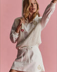 A woman holding a tennis racket poses against a pink background. She wears a relaxed silhouette featuring The Upside's Baccarat Violette Sweater, made from organic cotton, over a white shirt and an embroidered skirt.