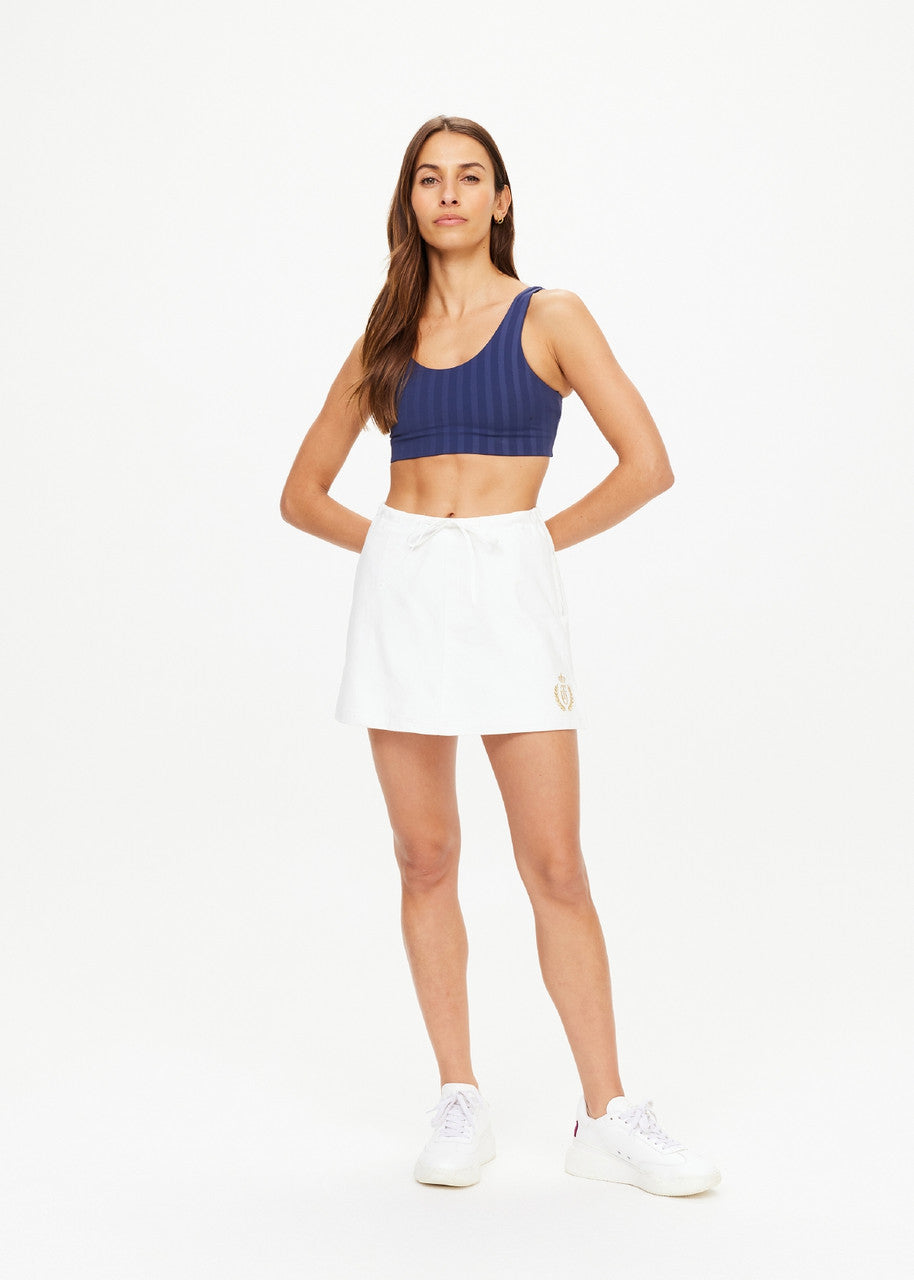 A woman poses confidently against a plain white background, showcasing her organic cotton navy sports bra and Baccarat Ruby Skort by The Upside. Her long hair is loose, and she completes the look with classic white sneakers, arms relaxed by her sides.