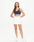 A woman poses confidently against a plain white background, showcasing her organic cotton navy sports bra and Baccarat Ruby Skort by The Upside. Her long hair is loose, and she completes the look with classic white sneakers, arms relaxed by her sides.