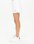 Side view of a person wearing the Baccarat Ruby Skort by The Upside, styled like classic tennis skorts, adorned with an emblem and paired with white sneakers. Made from organic cotton, this skort includes a small side slit. The individual is posed against a plain white background.