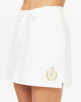 A person is wearing The Upside's Baccarat Ruby Skort, featuring a drawstring waist and crafted from organic cotton. This skort showcases a gold embroidered crest with a crown and laurel design near the hem, capturing the essence of classic tennis whites. The background is plain white.