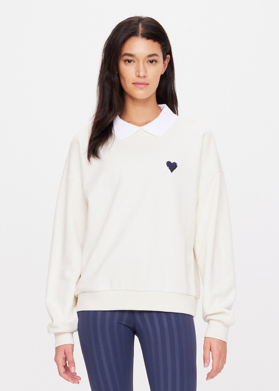 A person wearing The Upside's Pascal Collared Crew, made from organic cotton and adorned with a small embroidered heart logo on the left side. They pair it with blue striped pants, have long dark hair, and a neutral expression while standing against a plain background.