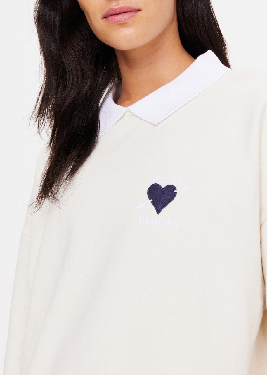 A person is wearing The Upside's Pascal Collared Crew, crafted from organic cotton in a cream hue. The chest features an elegantly embroidered navy blue heart with an arrow and the words "AGE OF HEARTS." The image focuses on the upper torso and part of the face, showcasing this distinctive embroidered logo.