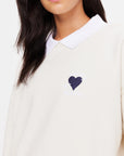 A person is wearing The Upside's Pascal Collared Crew, crafted from organic cotton in a cream hue. The chest features an elegantly embroidered navy blue heart with an arrow and the words "AGE OF HEARTS." The image focuses on the upper torso and part of the face, showcasing this distinctive embroidered logo.