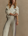 Against a beige backdrop, a person with long hair poses confidently, wearing The Upside's Pascal Collared Crew, an organic cotton cream-colored sweater featuring an embroidered heart emblem. Complementing the top are relaxed fit cream pants adorned with black, red, and blue stripes. Their hands on hips showcase a stylish array of jewelry.