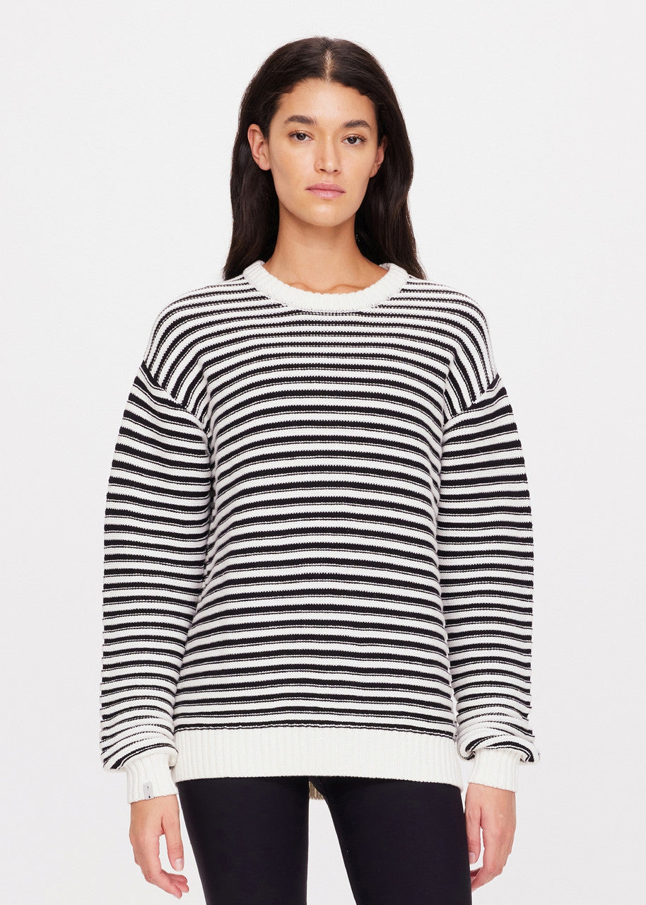 A person with long dark hair is wearing the Boo Knit Sweater by The Upside, a classic piece featuring black and white textured stripes, paired with dark pants. They stand against a plain white background, facing the camera.