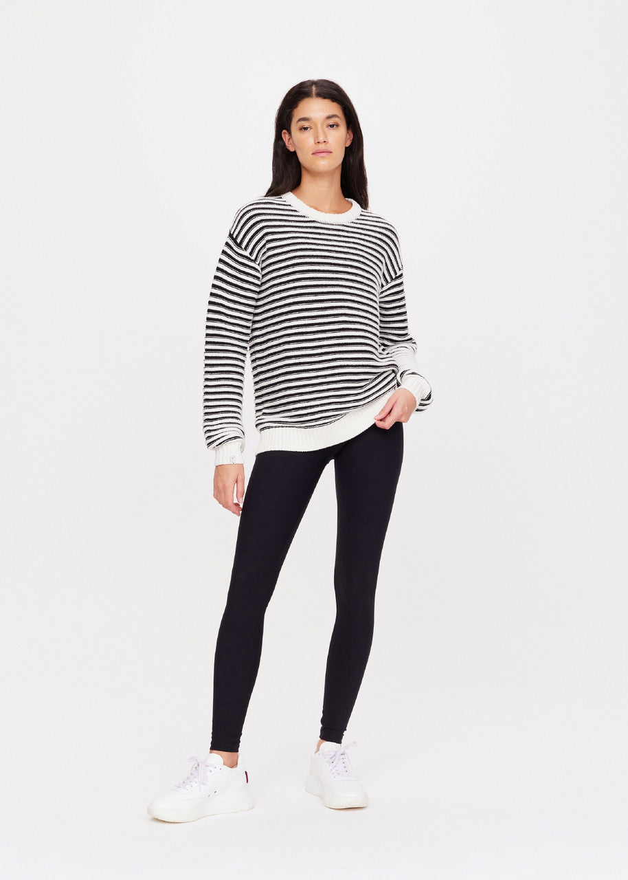 A person wearing The Upside's Boo Knit Sweater, paired with black leggings and white sneakers, stands against a plain white background. They have long dark hair and look directly at the camera with a relaxed posture.