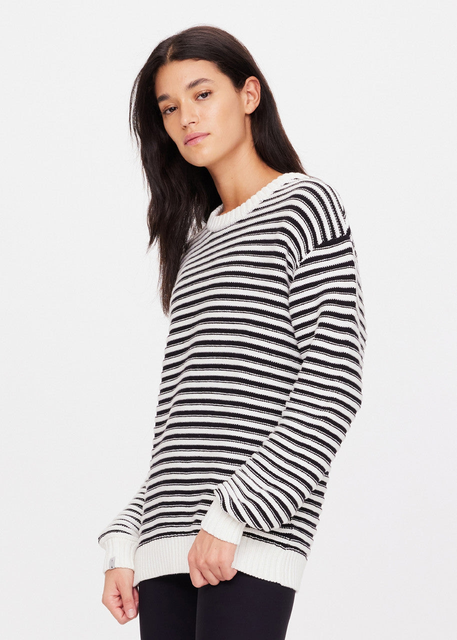 A person with long, dark hair wearing The Upside's Boo Knit Sweater stands against a plain white background. Crafted from organic cotton knit, the sweater's textured stripe pattern complements their neutral expression as they gaze slightly to the side.