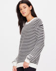 A person with long, dark hair wearing The Upside's Boo Knit Sweater stands against a plain white background. Crafted from organic cotton knit, the sweater's textured stripe pattern complements their neutral expression as they gaze slightly to the side.