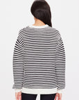 A person with long dark hair is wearing The Upside's Boo Knit Sweater, crafted from organic cotton, and standing with their back to the camera against a plain white background.