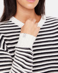 Dressed in The Upside's Boo Knit Sweater, featuring textured cream and black stripes, someone softly grasps the neckline. Crafted from organic combed cotton, its ribbed design elegantly contrasts against a pristine white backdrop.