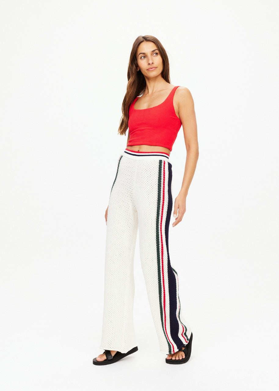 A woman in a vivid red sleeveless top and The Upside's Reverie Kansas Pant, featuring organic cotton with black, red, and white striped detailing down the sides, stands against a plain white background while gazing slightly to the side.