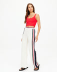 A woman in a vivid red sleeveless top and The Upside's Reverie Kansas Pant, featuring organic cotton with black, red, and white striped detailing down the sides, stands against a plain white background while gazing slightly to the side.