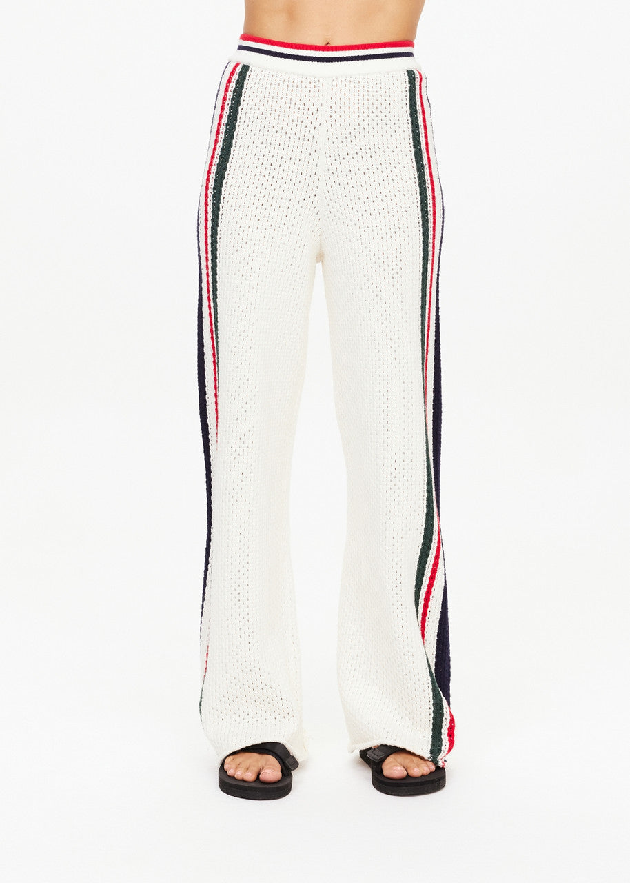 The Reverie Kansas Pant by The Upside features a white knit design with red, black, and white stripe detailing along the sides. Crafted from organic cotton, these wide-leg pants boast a high waist and are styled with sleek black slides against a minimalist white background.