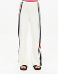 The Reverie Kansas Pant by The Upside features a white knit design with red, black, and white stripe detailing along the sides. Crafted from organic cotton, these wide-leg pants boast a high waist and are styled with sleek black slides against a minimalist white background.