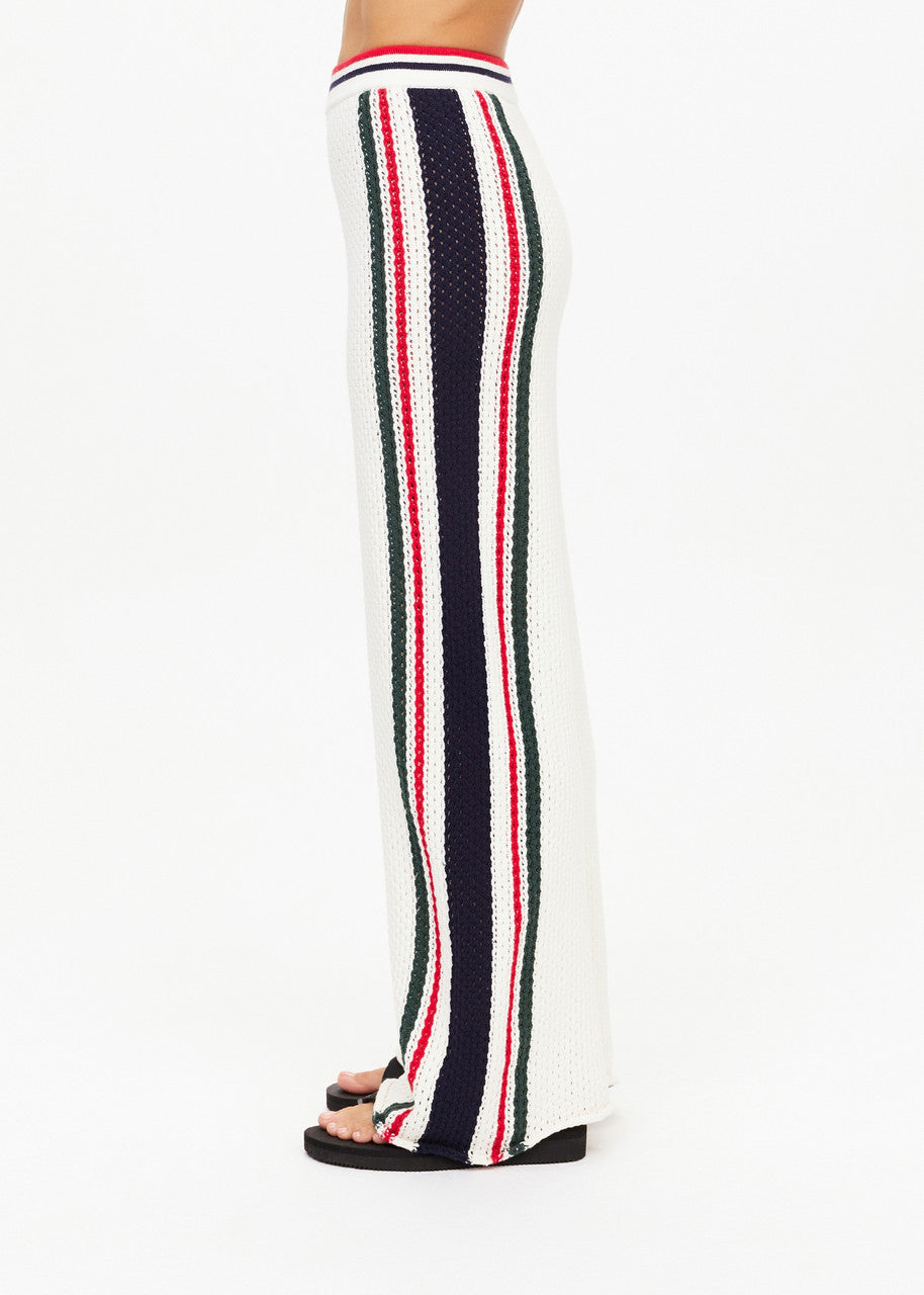 Side view of a person wearing the Reverie Kansas Pant by The Upside, featuring white crochet and a wide-leg silhouette with a navy, red, and green striped knit design. Made from organic cotton, these sporty retro pants pair effortlessly with black sandals against a neutral background.