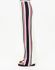 Side view of a person wearing the Reverie Kansas Pant by The Upside, featuring white crochet and a wide-leg silhouette with a navy, red, and green striped knit design. Made from organic cotton, these sporty retro pants pair effortlessly with black sandals against a neutral background.