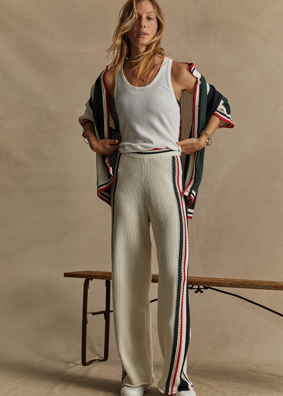 A person wearing a white tank top and The Upside's Reverie Kansas Pant, featuring an organic cotton blend with a wide-leg silhouette highlighted by red and green stripes, stands indoors on a wooden bench. They have a striped cardigan draped over their shoulders against a beige background.