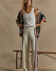 A person wearing a white tank top and The Upside's Reverie Kansas Pant, featuring an organic cotton blend with a wide-leg silhouette highlighted by red and green stripes, stands indoors on a wooden bench. They have a striped cardigan draped over their shoulders against a beige background.