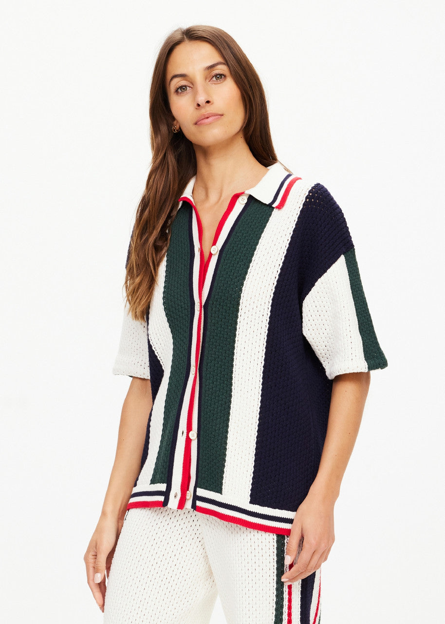 A woman with long brown hair is modeling a Reverie Chino Shirt from The Upside. This short-sleeved, open-knit cardigan, crafted from organic cotton, showcases vertical stripes in navy, green, white, and red. She complements her look with coordinated knit pants against a plain white background.