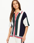 A woman with long brown hair is modeling a Reverie Chino Shirt from The Upside. This short-sleeved, open-knit cardigan, crafted from organic cotton, showcases vertical stripes in navy, green, white, and red. She complements her look with coordinated knit pants against a plain white background.