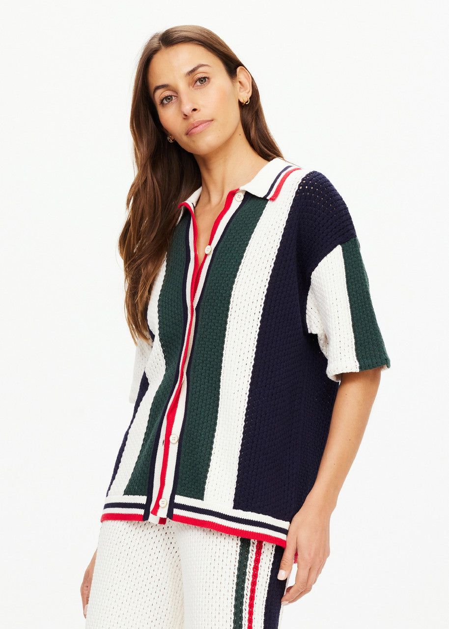 A woman wearing The Upside's Reverie Chino Shirt and matching pants poses against a white background. The relaxed-fit outfit showcases vertical navy, green, and white stripes with retro red accents. Made from organic cotton, she has long brown hair and a neutral expression.