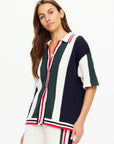 A woman wearing The Upside's Reverie Chino Shirt and matching pants poses against a white background. The relaxed-fit outfit showcases vertical navy, green, and white stripes with retro red accents. Made from organic cotton, she has long brown hair and a neutral expression.