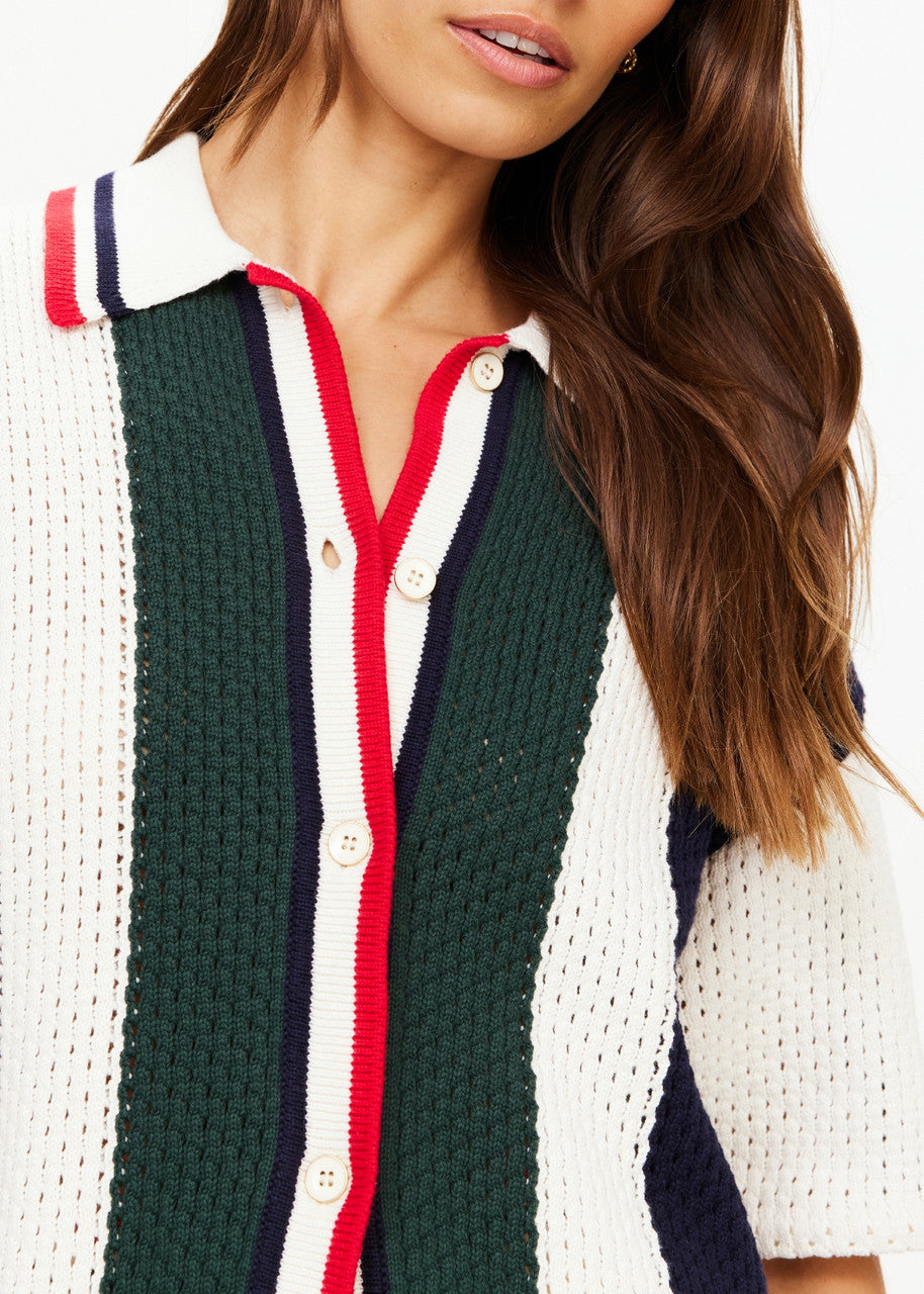 A person wearing The Upside's Reverie Chino Shirt in a retro style featuring green, white, and navy color-blocked knit with red and navy trim and buttons. Made from organic cotton, the shirt showcases a wide collar and short sleeves. Long brown hair cascades down one side as the image focuses on the upper torso.