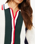 A person wearing The Upside's Reverie Chino Shirt in a retro style featuring green, white, and navy color-blocked knit with red and navy trim and buttons. Made from organic cotton, the shirt showcases a wide collar and short sleeves. Long brown hair cascades down one side as the image focuses on the upper torso.