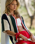 A person with long hair leans against a post, wearing the Reverie Chino Shirt by The Upside over a white crop top and red shorts. They hold a tennis racket and have an organic cotton bandana wrapped around their wrist, maintaining a neutral expression outdoors.