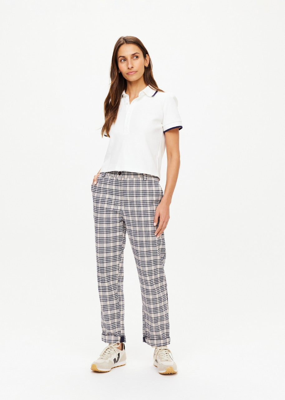 A person with long hair is wearing a white polo shirt and The Upside's Highlands Pant, featuring a plaid design and an elastic waist. They stand against a plain white background, looking slightly to the right, with one hand in their pocket and are wearing white sneakers.