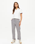 A person with long hair is wearing a white polo shirt and The Upside's Highlands Pant, featuring a plaid design and an elastic waist. They stand against a plain white background, looking slightly to the right, with one hand in their pocket and are wearing white sneakers.