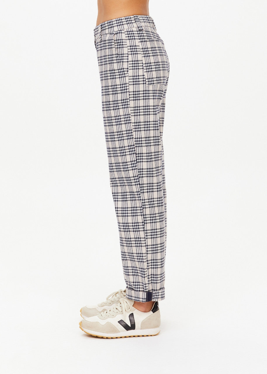 A side profile showcases a person wearing The Upside's Highlands Pant in a stylish white and navy check pattern. These pants, crafted from organic cotton, are designed with an elastic waist for both comfort and style. They are paired with sleek white sneakers against a plain white backdrop.