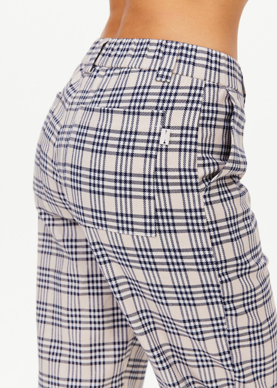 A close-up image showcases someone dressed in The Upside Highlands Pant, a beige and black plaid garment. These pants feature an elastic waist and a classic checkered pattern crafted from organic cotton. There is also a back pocket with a small label visible. The background is plain white.