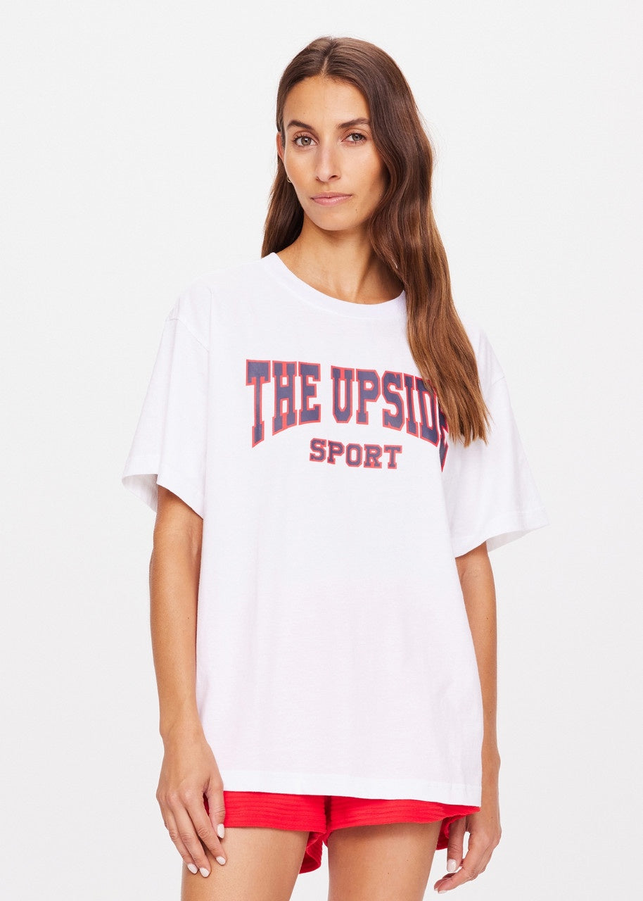 A person with long brown hair is wearing an oversized Ivy League Sam Tee made from organic cotton, emblazoned with "The Upside" in red and black paired with red shorts. They stand against a plain white background.