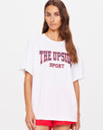 A person with long brown hair is wearing an oversized Ivy League Sam Tee made from organic cotton, emblazoned with "The Upside" in red and black paired with red shorts. They stand against a plain white background.