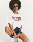 A person with curly hair wearing sunglasses and an oversized Ivy League Sam Tee by The Upside in organic cotton is paired with blue shorts and white sneakers, kneeling on one knee. They enhance the outfit with necklaces, a bracelet, and a blue bandana tied around their wrist.