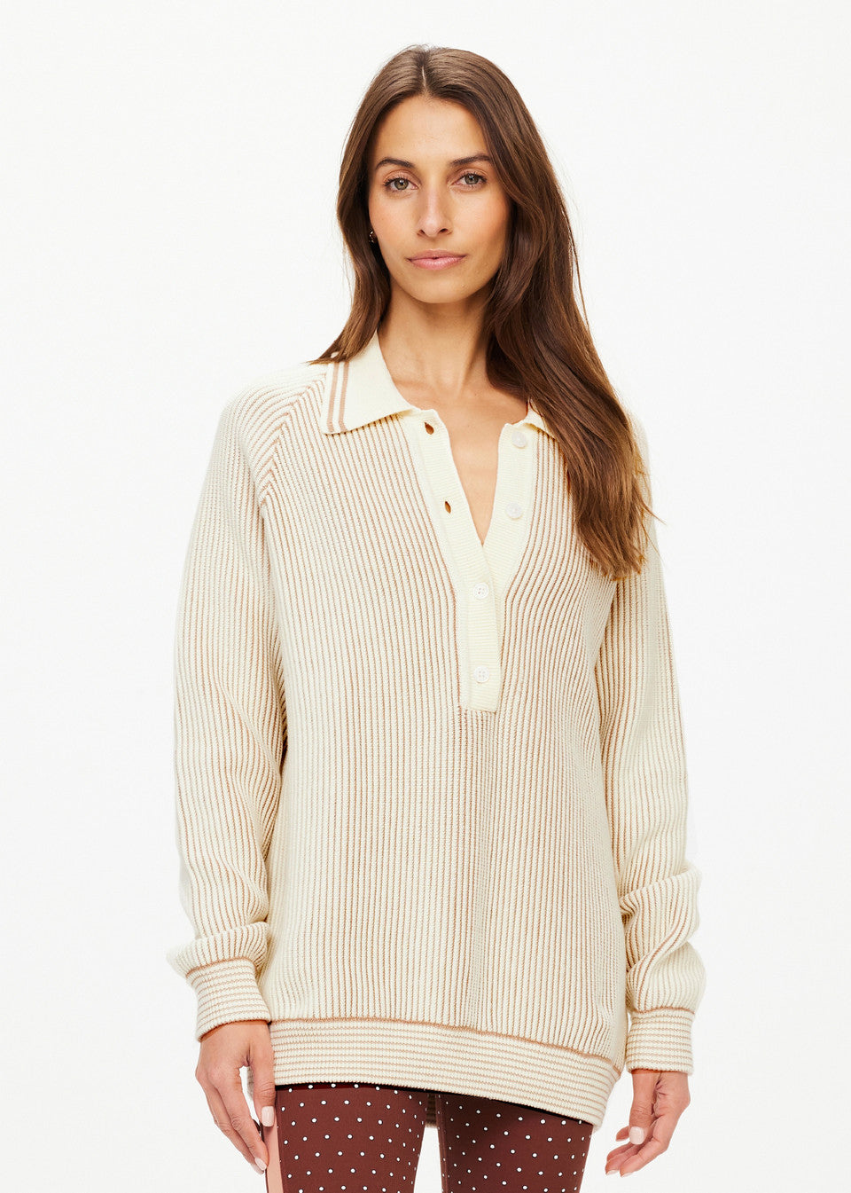 A person with long hair is wearing The Upside's ALANA KNITTED SWEATER, a cream-colored ribbed design featuring a collar and buttons. They have paired it with brown polka dot pants, standing against a plain white background.