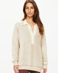 A person with long hair is wearing The Upside's ALANA KNITTED SWEATER, a cream-colored ribbed design featuring a collar and buttons. They have paired it with brown polka dot pants, standing against a plain white background.