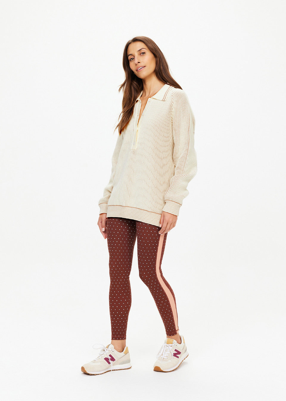 In a white setting, someone wearing The Upside's ALANA KNITTED SWEATER and brown polka dot leggings stands comfortably. They have long brown hair and sport white sneakers with red touches.