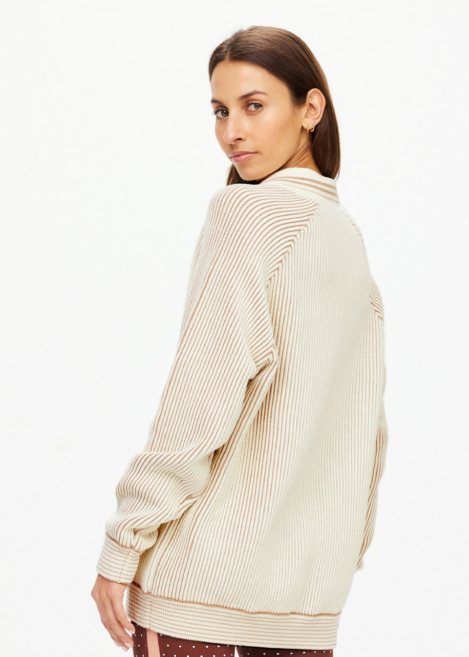 A woman with long brown hair wears The Upside's ALANA KNITTED SWEATER, cream-colored and striped. She stands against a plain white background, glancing over her shoulder with a slight smile.