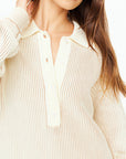 A person in The Upside's cream ALANA KNITTED SWEATER, featuring a textured knit with a collar and button-up design, displays vertical ribbing. One hand touches the neck, while their long, slightly wavy brown hair cascades down.