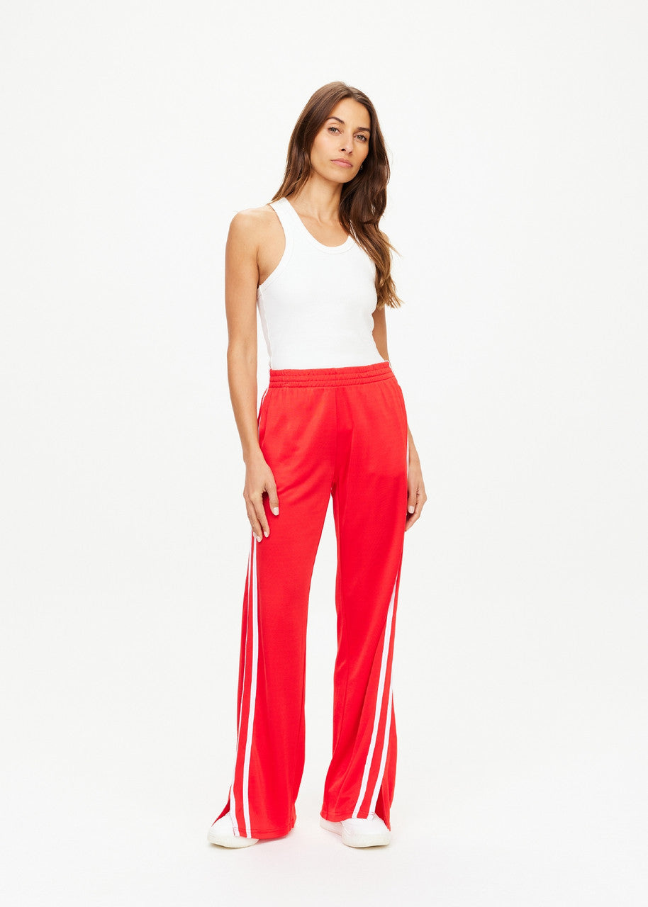 Against a plain backdrop, an individual with long hair confidently showcases their style by wearing a white tank top along with The Upside's Juliet Pant in red, featuring white stripes. These low-rise pants are crafted from slinky jersey fabric and pair seamlessly with both their white shoes and relaxed demeanor.