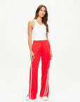 Against a plain backdrop, an individual with long hair confidently showcases their style by wearing a white tank top along with The Upside's Juliet Pant in red, featuring white stripes. These low-rise pants are crafted from slinky jersey fabric and pair seamlessly with both their white shoes and relaxed demeanor.