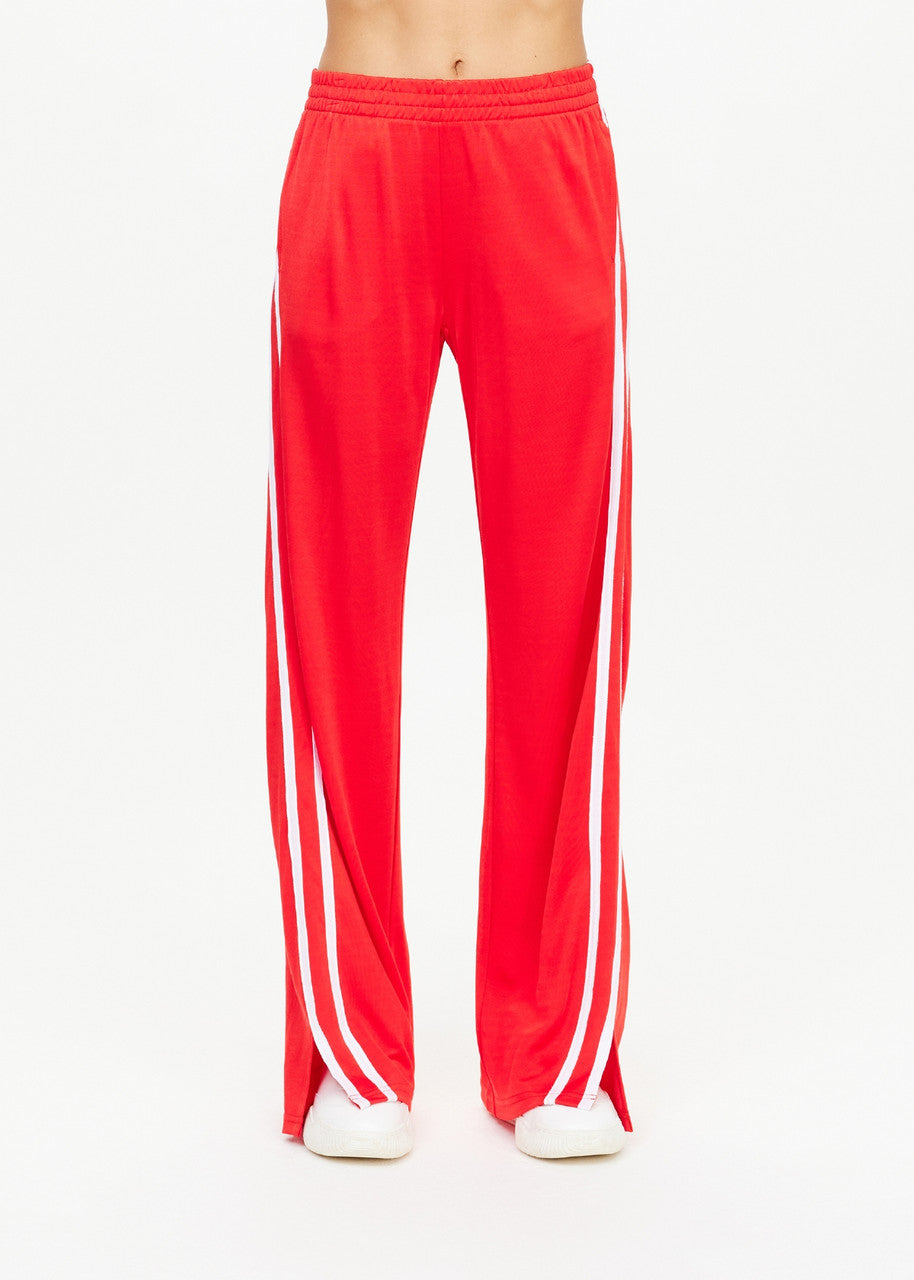 A person dressed in The Upside's Juliet Pant track pants, featuring a vibrant red color with white side stripes and made from a sleek double-faced jersey, stands against a simple backdrop. Their midsection is visible, and they are showcasing trendy white shoes.