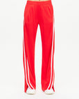 A person dressed in The Upside's Juliet Pant track pants, featuring a vibrant red color with white side stripes and made from a sleek double-faced jersey, stands against a simple backdrop. Their midsection is visible, and they are showcasing trendy white shoes.