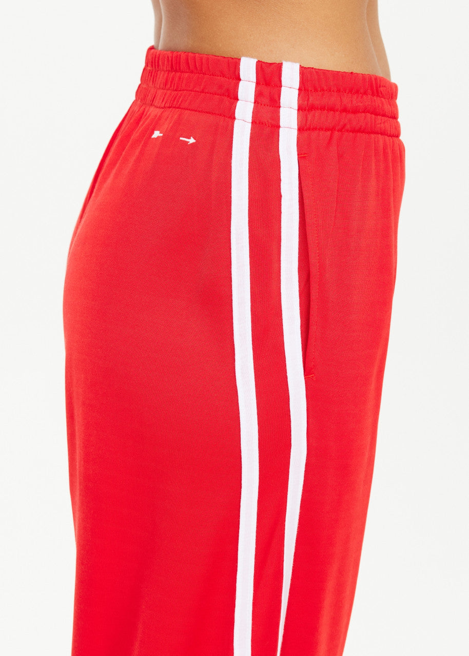 A person is wearing The Upside's Juliet Pant, which is red with white side stripes. These low-rise pants are made from a soft double-faced jersey and include an elastic waistband, along with a small white logo near the hip. The background is plain white.